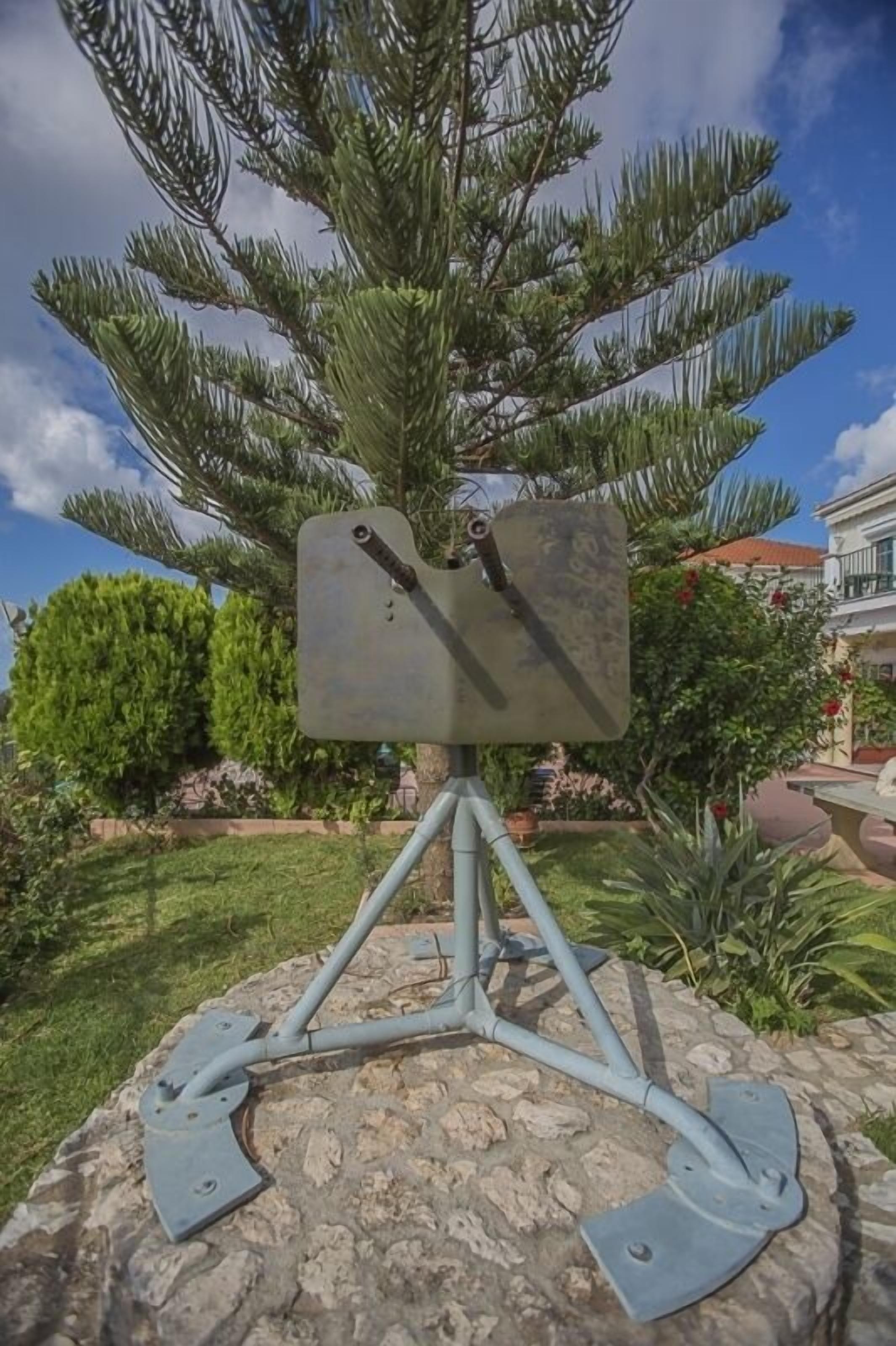 Un panneau métallique, de forme rectangulaire, fixé sur un trépied et placé sur un socle en pierre, dans un jardin agrémenté d’un pin et d’autres plantes vertes en arrière-plan.