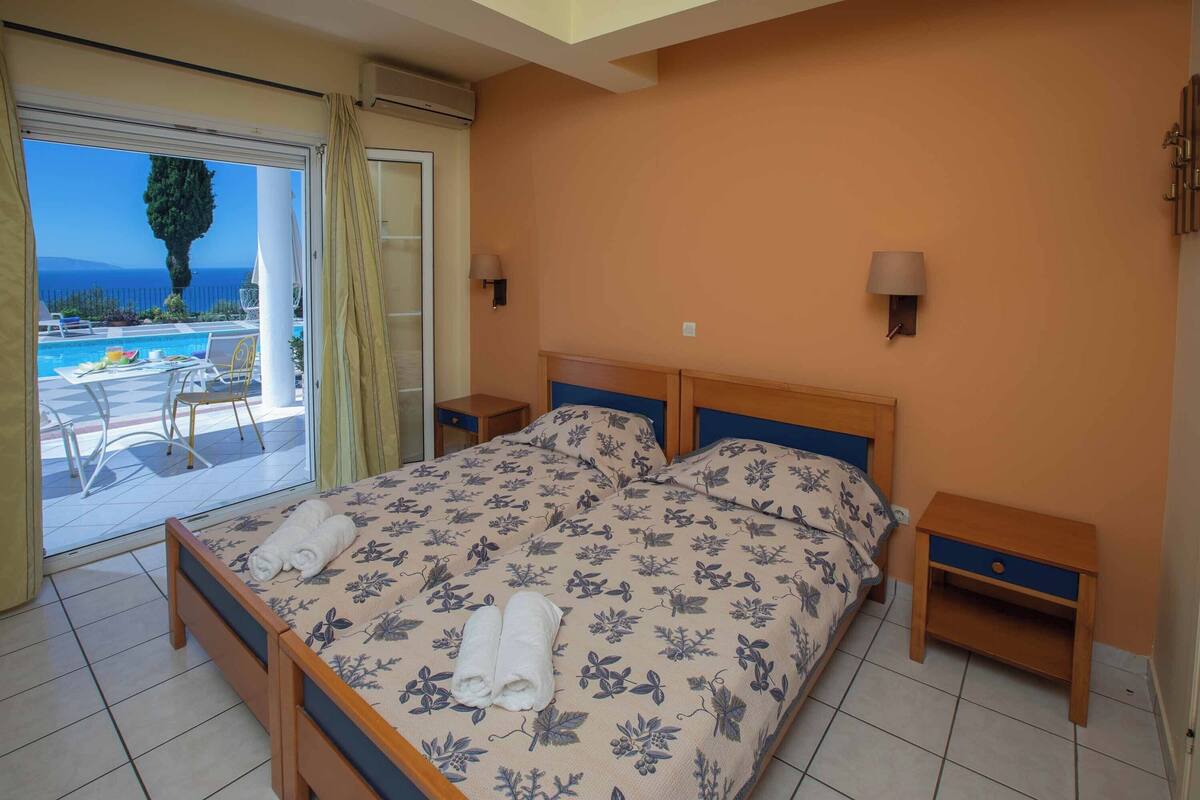 A bedroom with a bed, two nightstands, a window with curtains, and a view of a pool and landscape.