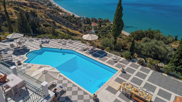 A pool area with checkered flooring, lounge chairs, and umbrellas, overlooking a coastal landscape with a beach and hills. - Garbis Villas (Kefalonia)