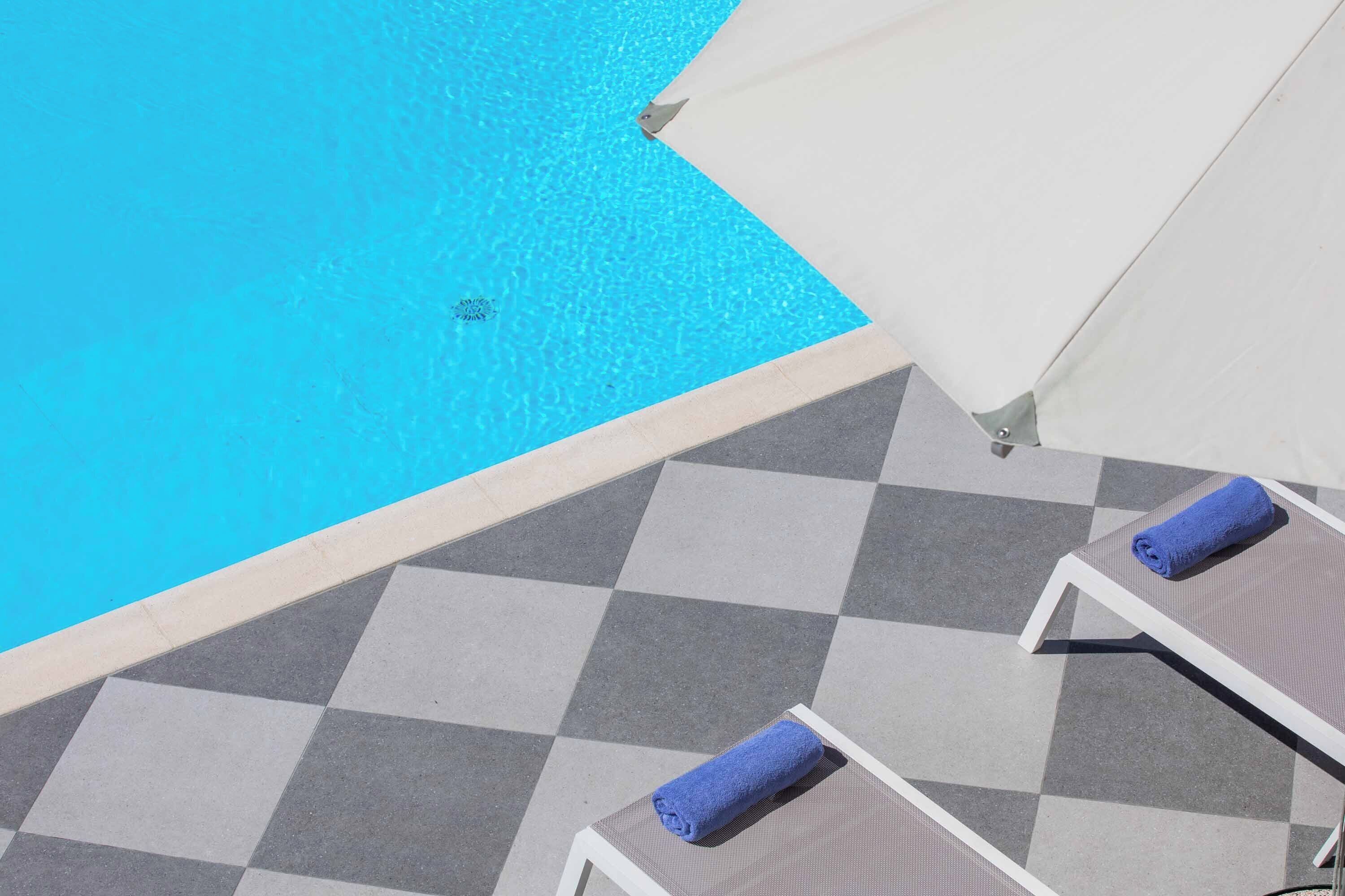 Un área junto a la piscina con piso a cuadros, sillas de descanso con toallas azules y un paraguas blanco.