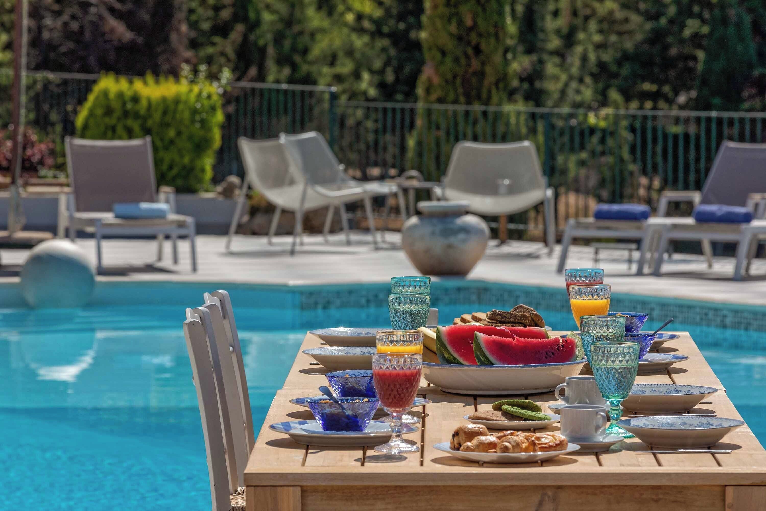 A poolside dining table with plates, glasses, and a watermelon.