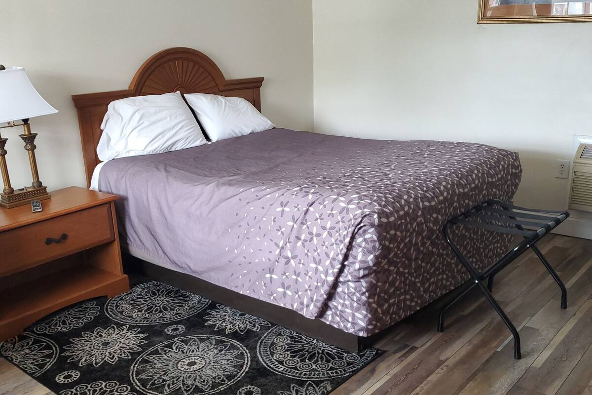 A neatly arranged bedroom with a bed, a nightstand, a lamp, a patterned rug, and a wall-mounted air conditioner.