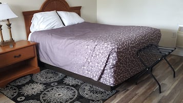 A neatly arranged bedroom with a bed, a nightstand, a lamp, a patterned rug, and a wall-mounted air conditioner.