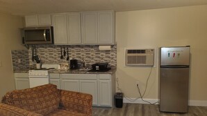 Una cocina compacta con armarios blancos, un microondas, una estufa y un refrigerador. Un sofá con diseño destaca al frente.