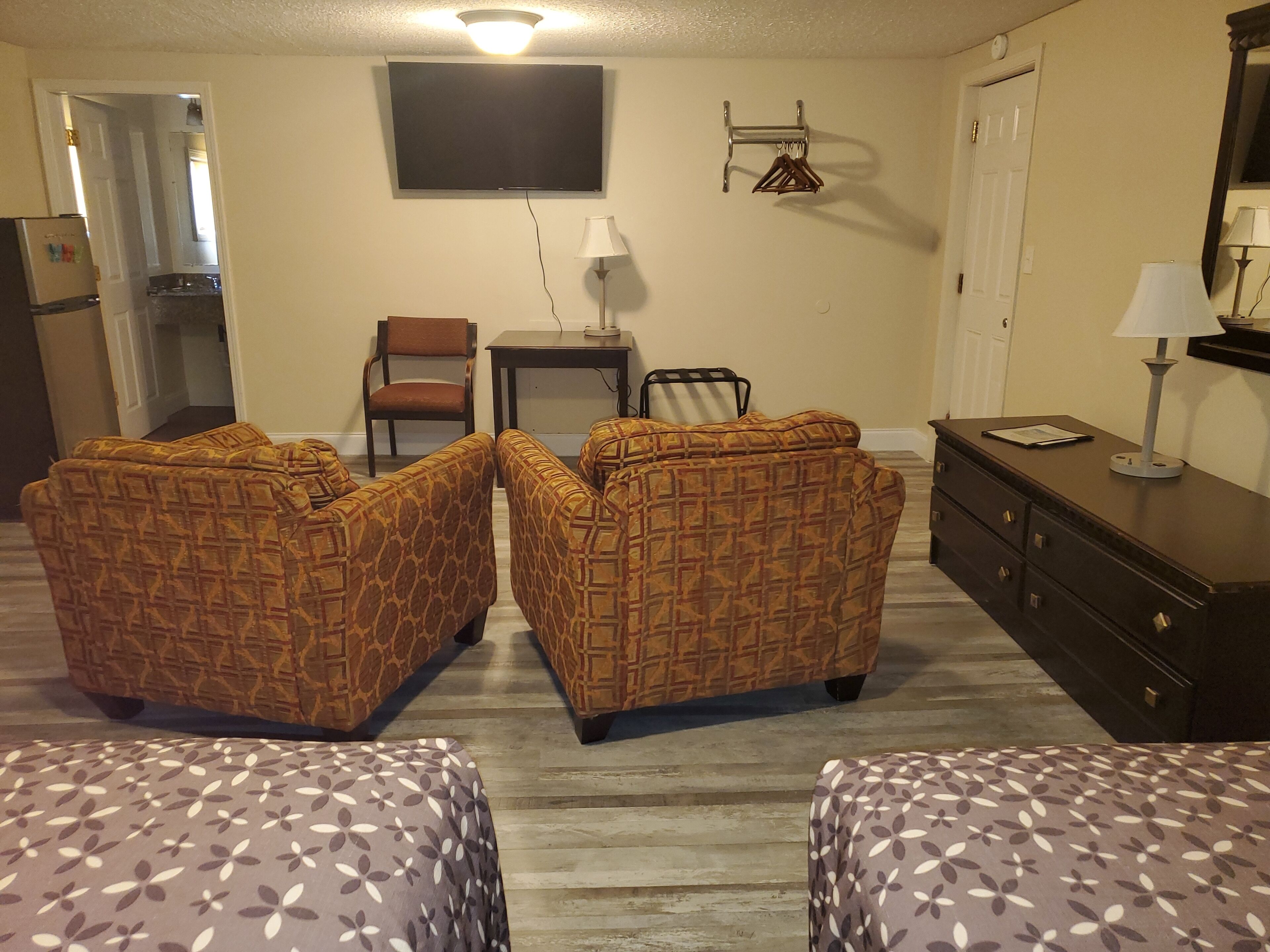 A room with two patterned sofas, a small table, a chair, a dresser, and a television on the wall.