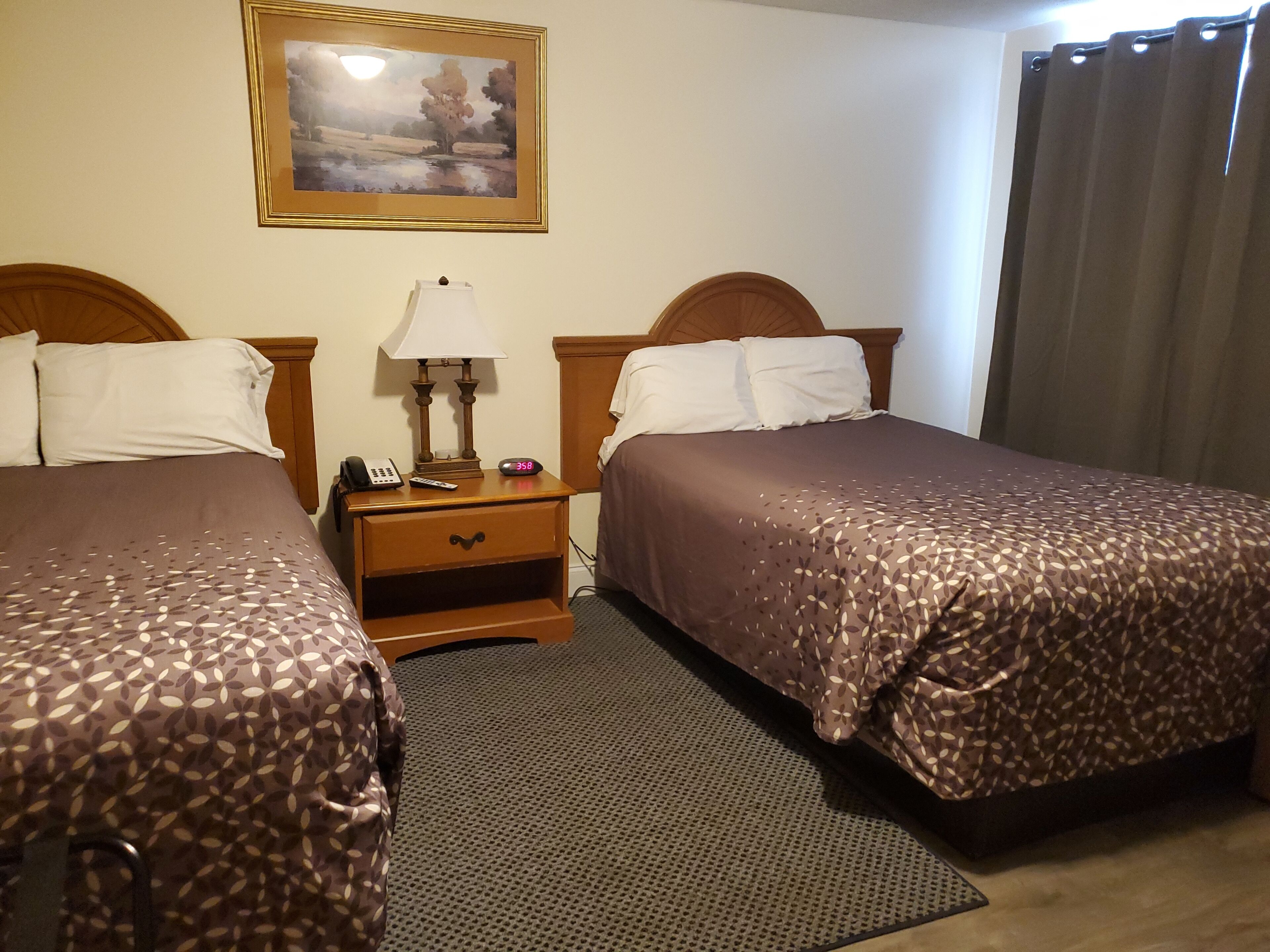 A hotel room with two beds, a nightstand with a lamp, a painting on the wall, and a window with curtains.