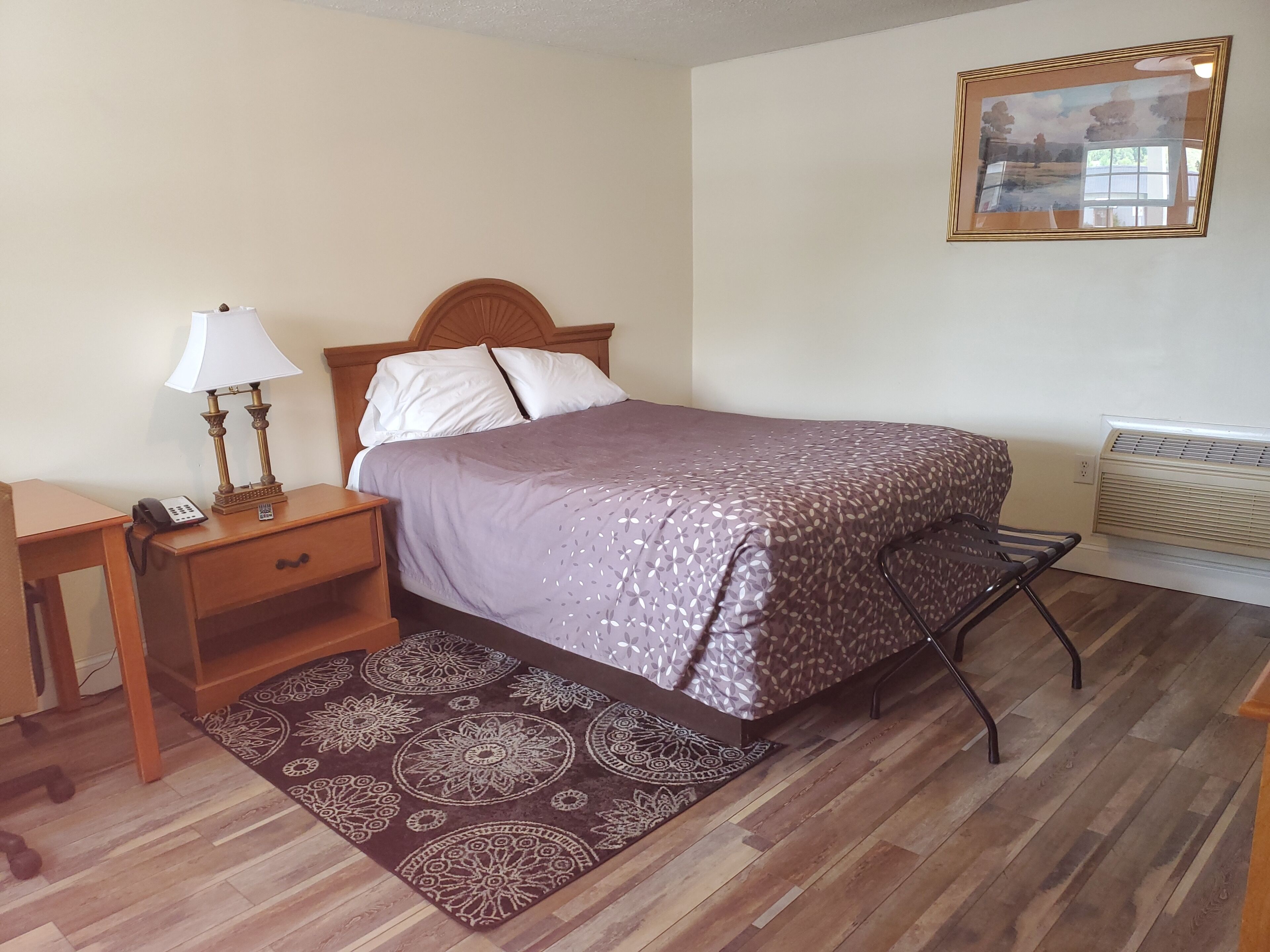 A neatly arranged bedroom with a bed, bedside table with a lamp, a chair, a rug, and a wall-mounted air conditioner.