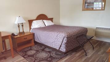 A neatly arranged bedroom with a bed, bedside table with a lamp, a chair, a rug, and a wall-mounted air conditioner.