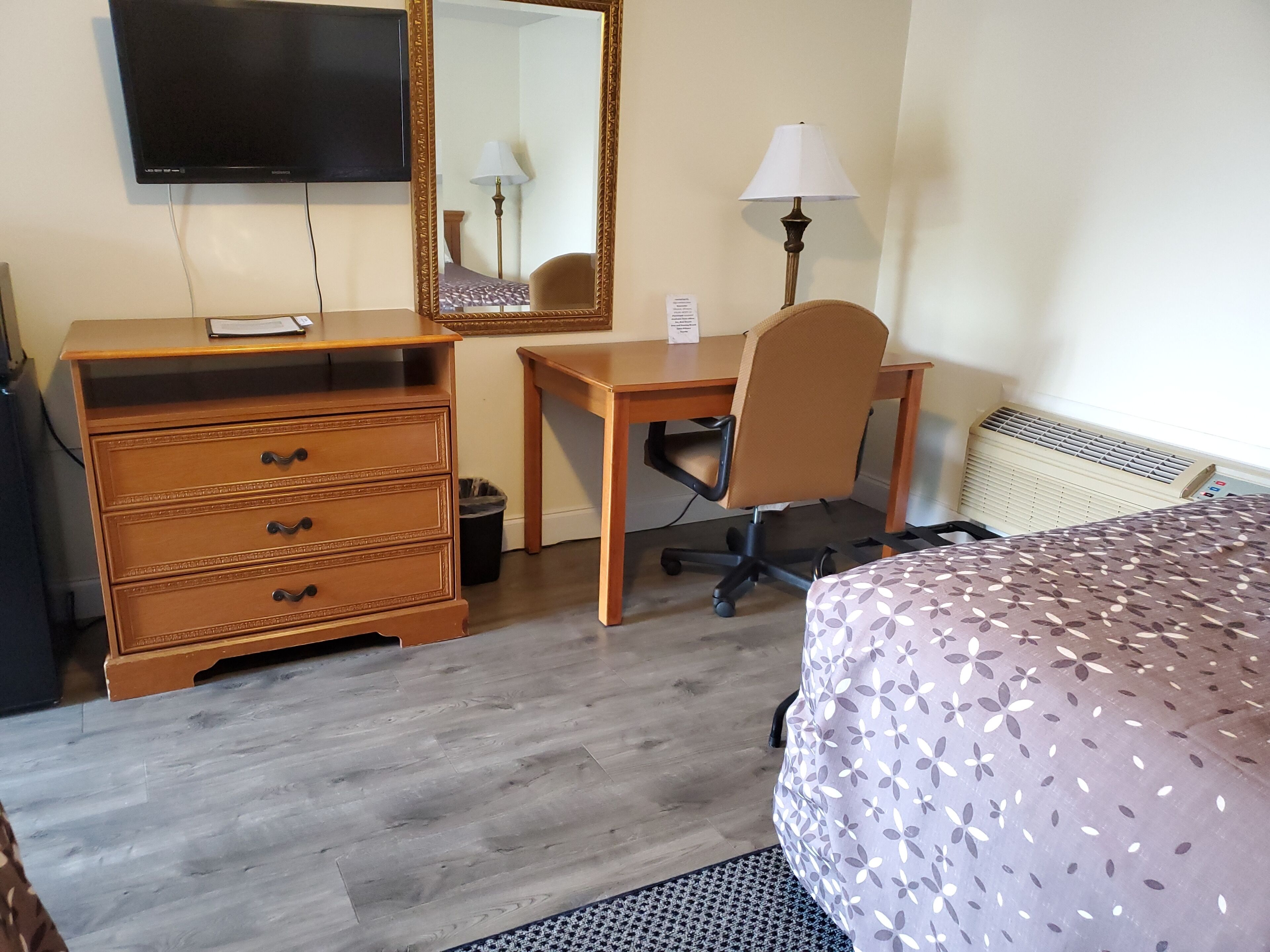 A hotel room with a wooden desk, a chair, a dresser, a bed with a patterned cover, a television, a mirror, and a lamp.