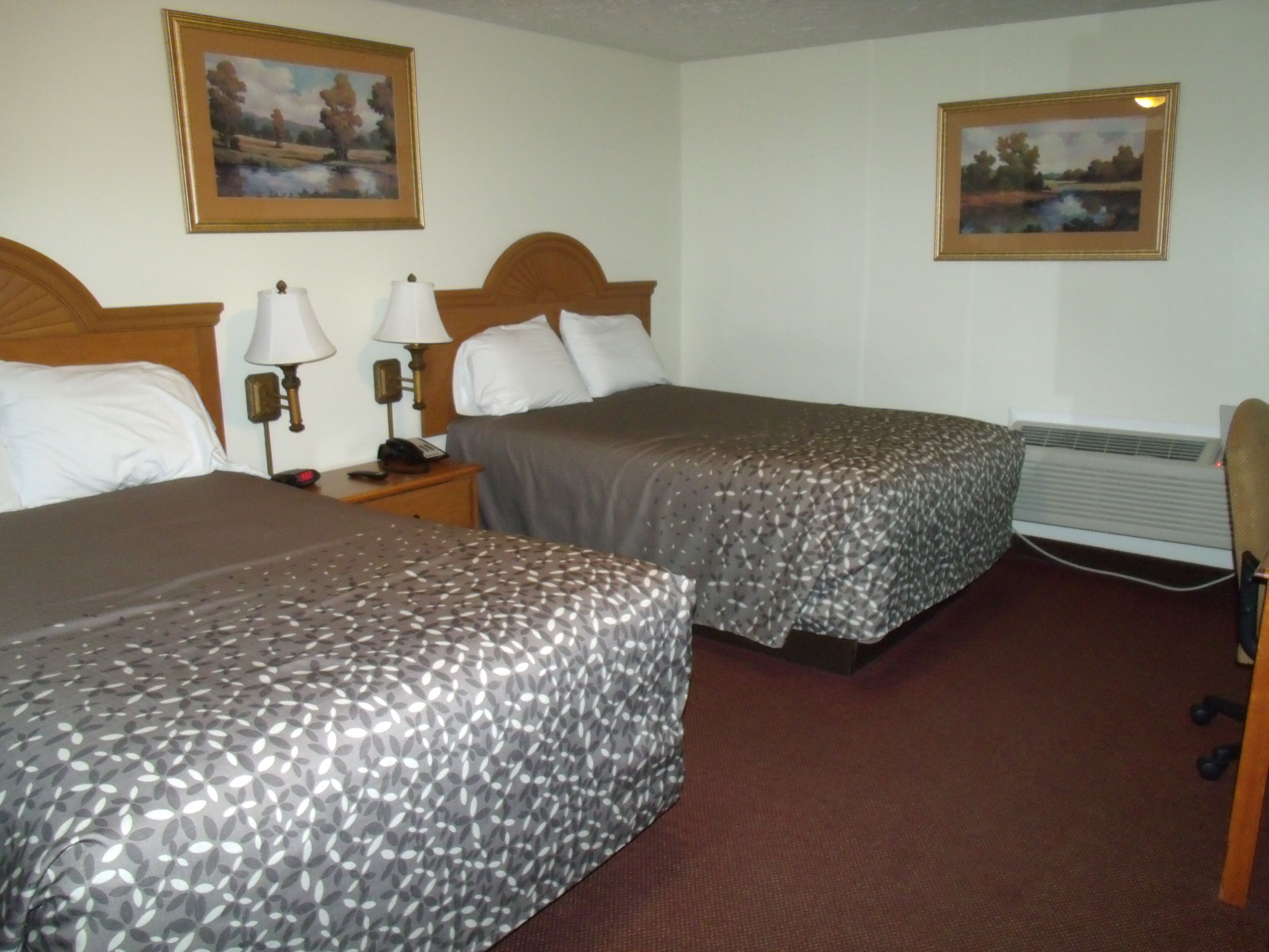 A hotel room with two beds, a nightstand with a lamp, a telephone, and an air conditioning unit.