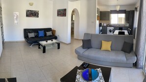A modern living room with a grey sofa, a black coffee table, and a glass coffee table. There is a kitchen visible in the background with a washing machine and a dining area with a table and chairs. - Sirena Sunrise (Paphos)
