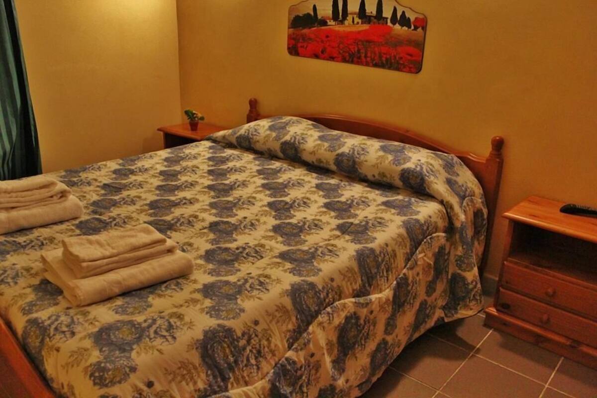 Un lit bien fait, recouvert d’une couvre-lit à motifs floraux, une petite table de chevet avec une plante et un tableau accroché au mur.