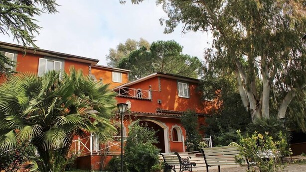 A two-story building with a red facade, a covered entrance, and a small garden area with benches and palm trees. - Agriturismo Marco Simone (Rome)