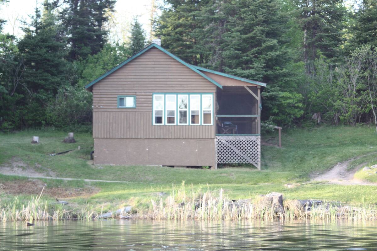 Une petite cabane avec un porche, située au bord d’un lac, entourée d’arbres.