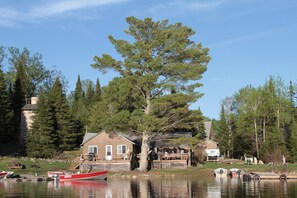 Järven rannalla sijaitseva mökki, jossa on laituri, ja sen ympärillä on puita sekä pieni vene.