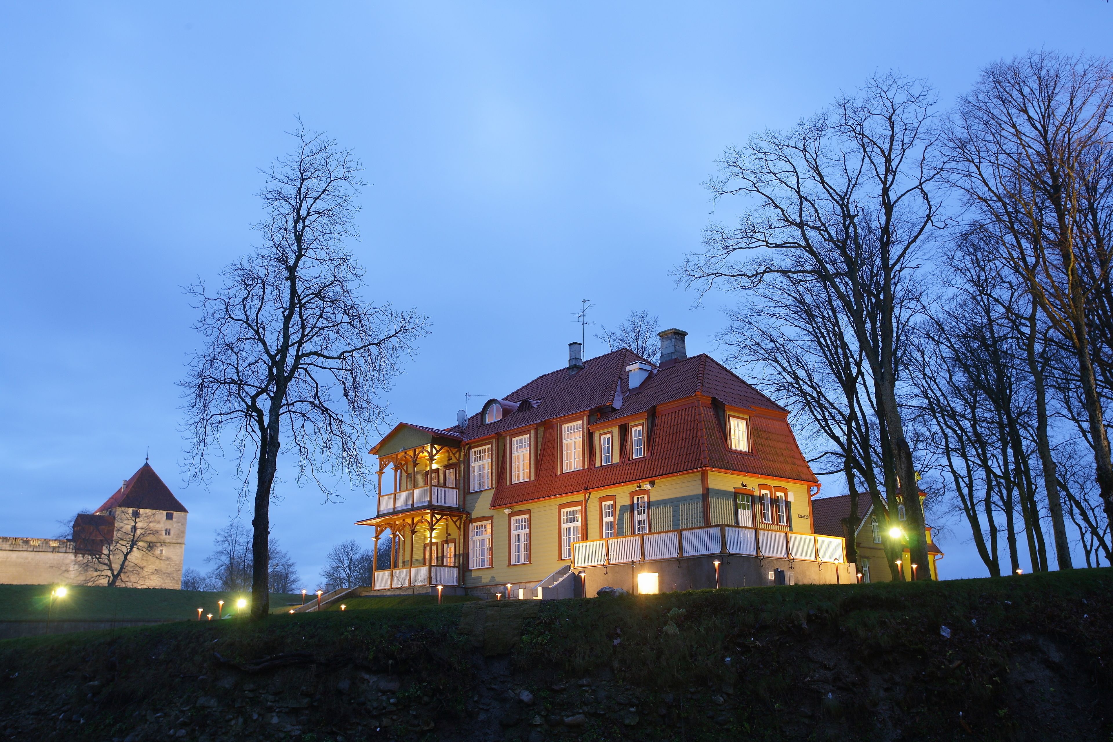 A large, multi-story building with a red roof and multiple windows, surrounded by trees and a stone structure in the background.