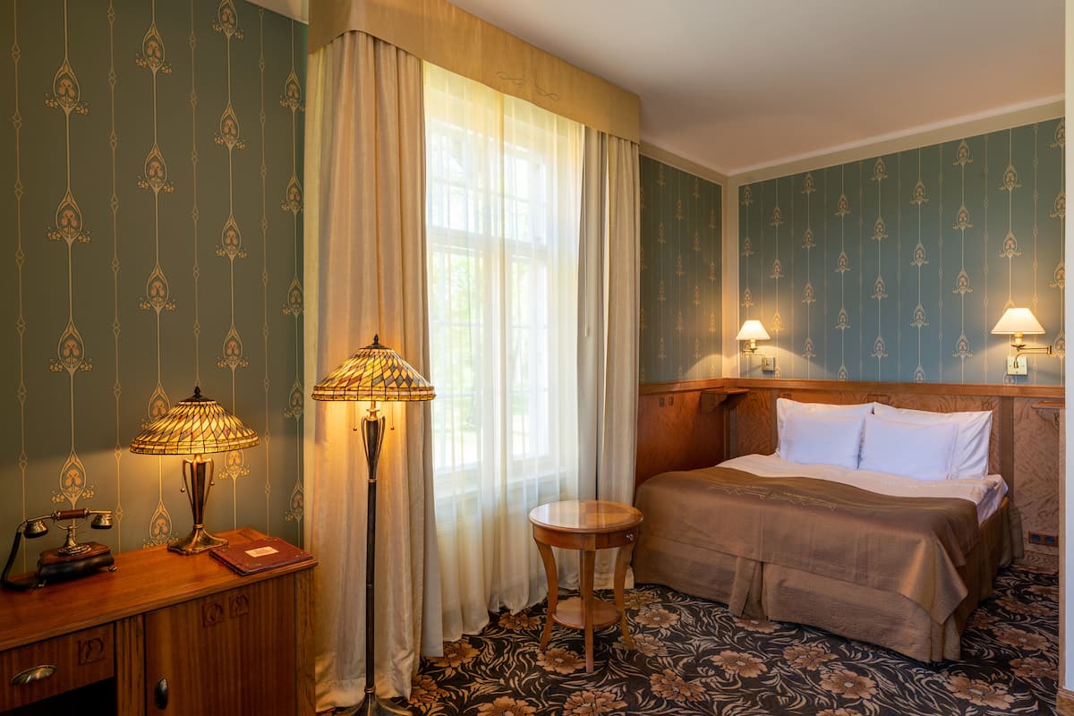 A hotel room with a bed, a desk with a lamp, a small table, and curtains.