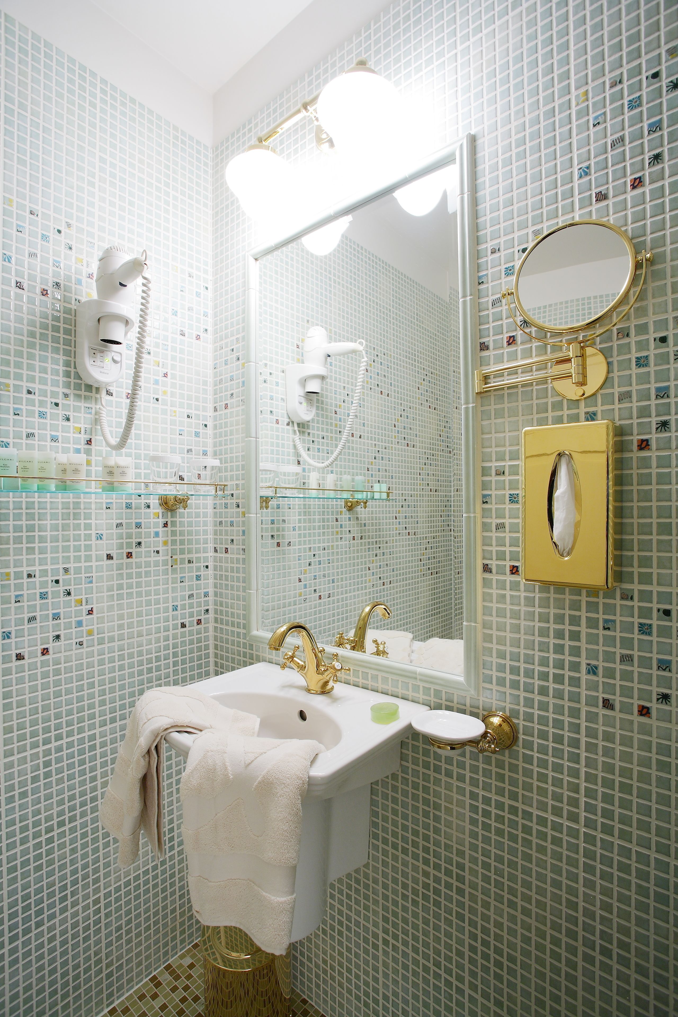 Un baño con lavamanos blanco, detalles dorados, un espejo y un perchero.