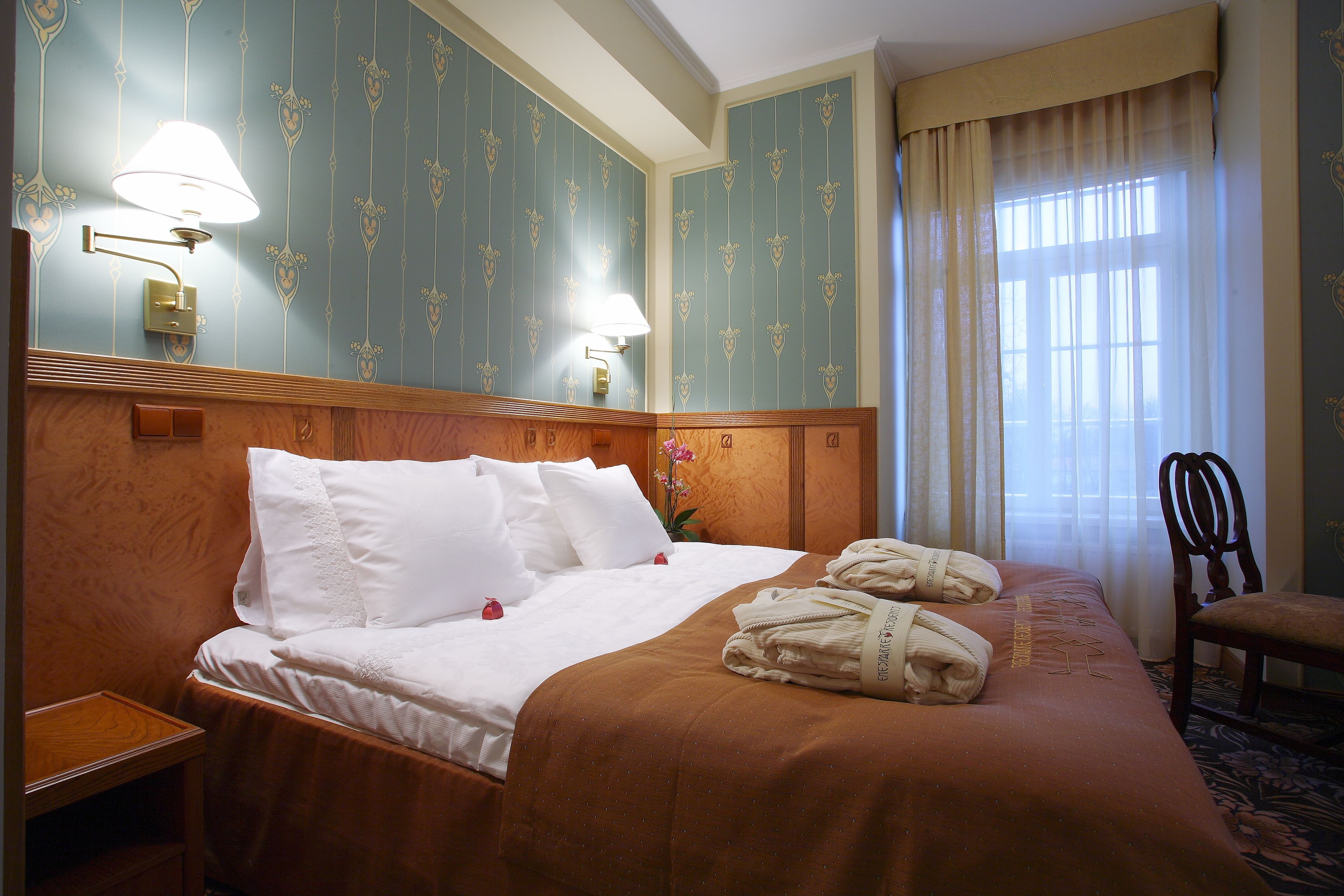 A hotel room with a neatly made bed, a wooden nightstand, a chair, and a window with curtains.