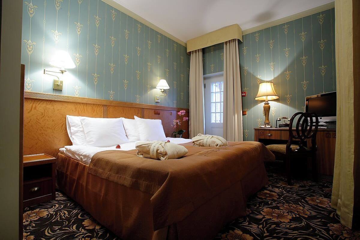 A hotel room with a double bed, two bedside lamps, a wooden headboard, a desk with a chair, and a patterned carpet.