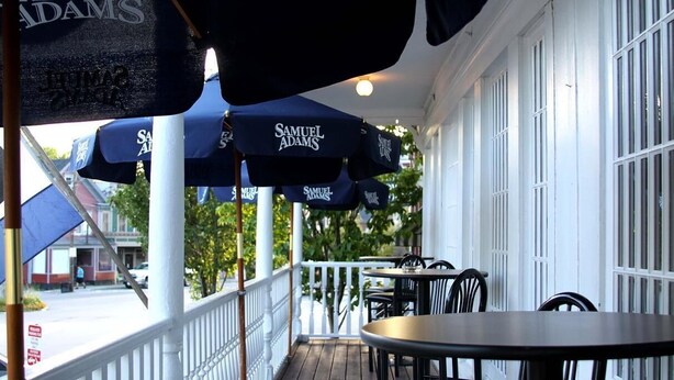 A covered outdoor seating area with tables and chairs, umbrellas with 'Samuel Adams' branding, and a white railing. - Tilton Inn (Tilton)
