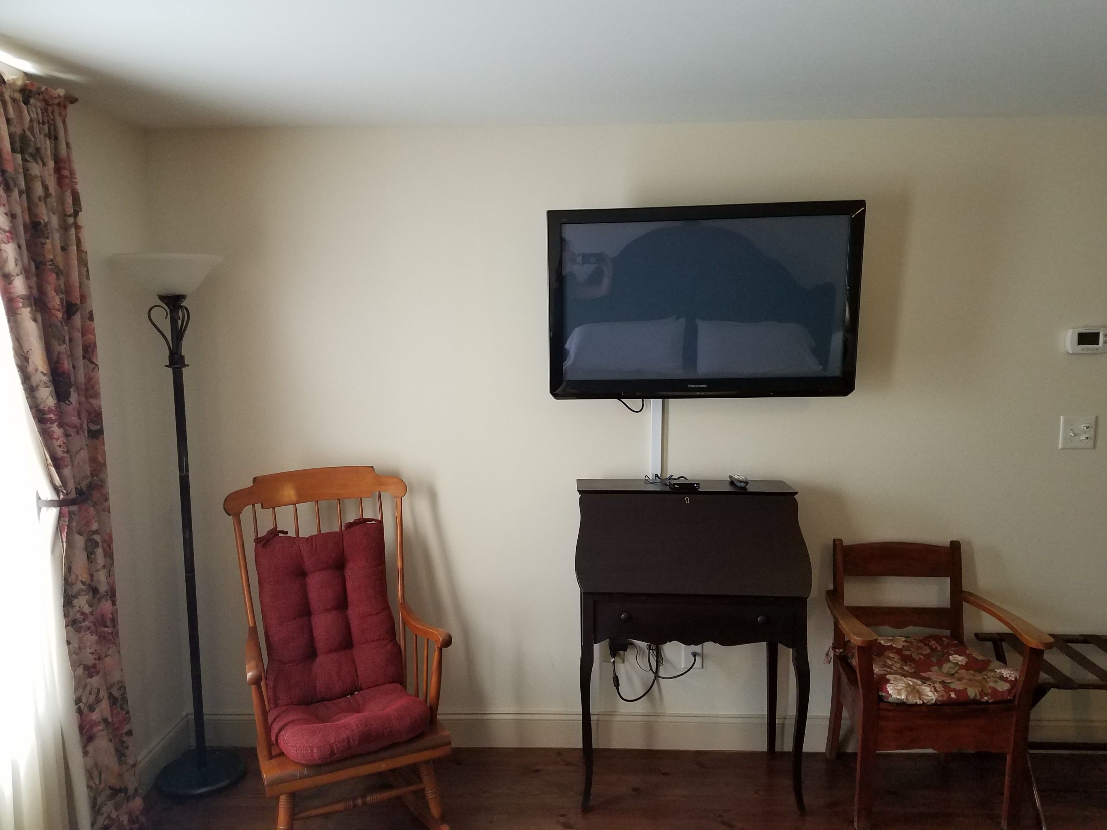 A living room with a television mounted on the wall, a wooden chair with a cushion, and a floor lamp.