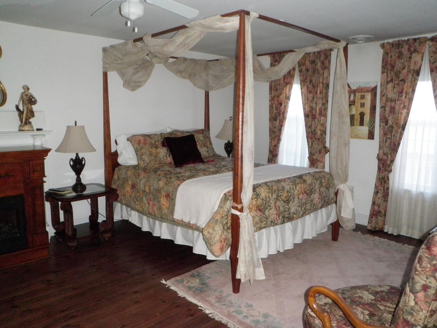 A four-poster canopy bed with floral bedding, a wooden nightstand with a lamp, and a chair with floral upholstery.