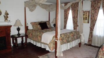 A four-poster canopy bed with floral bedding, a wooden nightstand with a lamp, and a chair with floral upholstery.