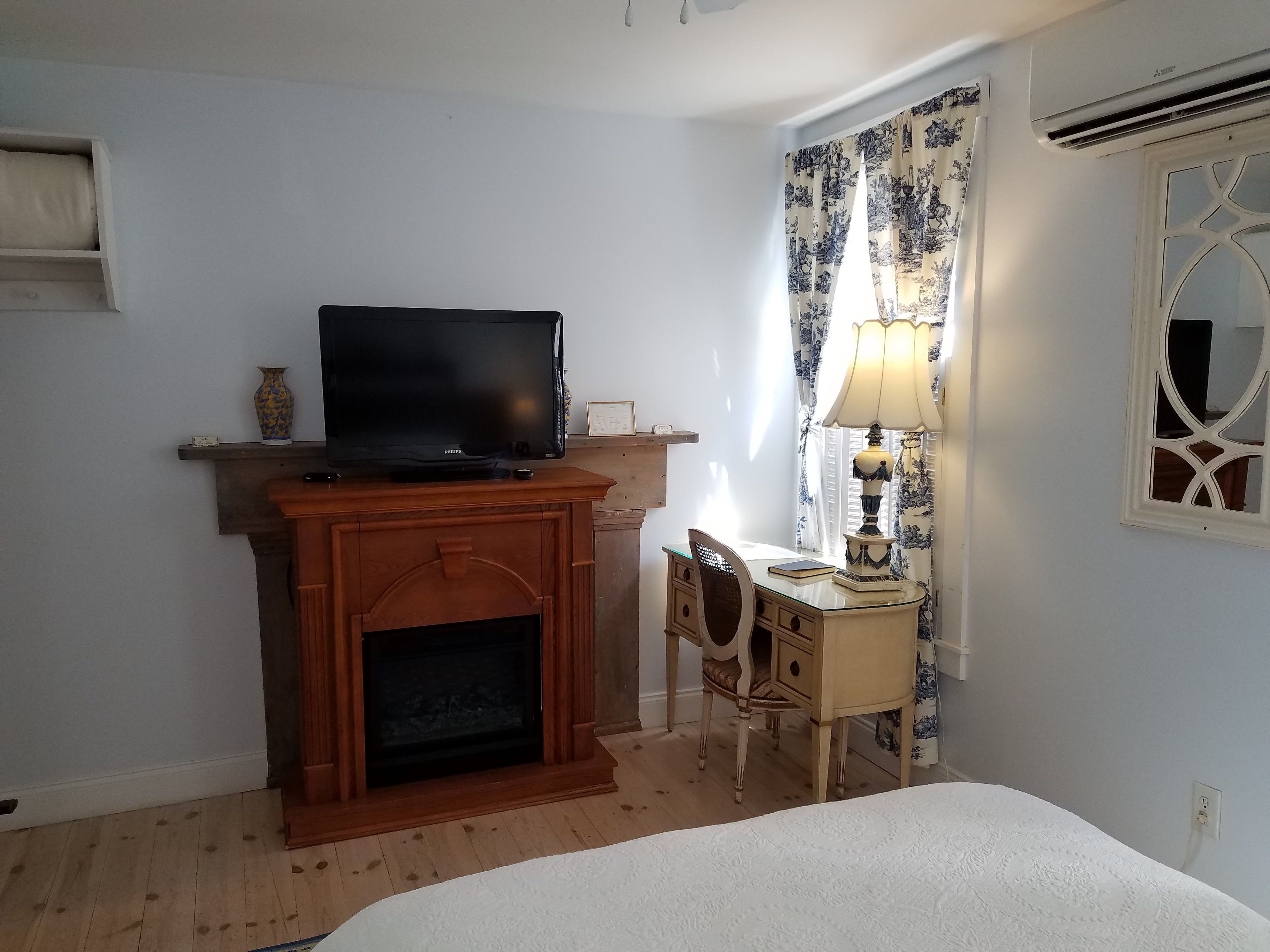 A hotel room with a television on a wooden stand, a desk with a lamp, and a bed with a white bedspread.
