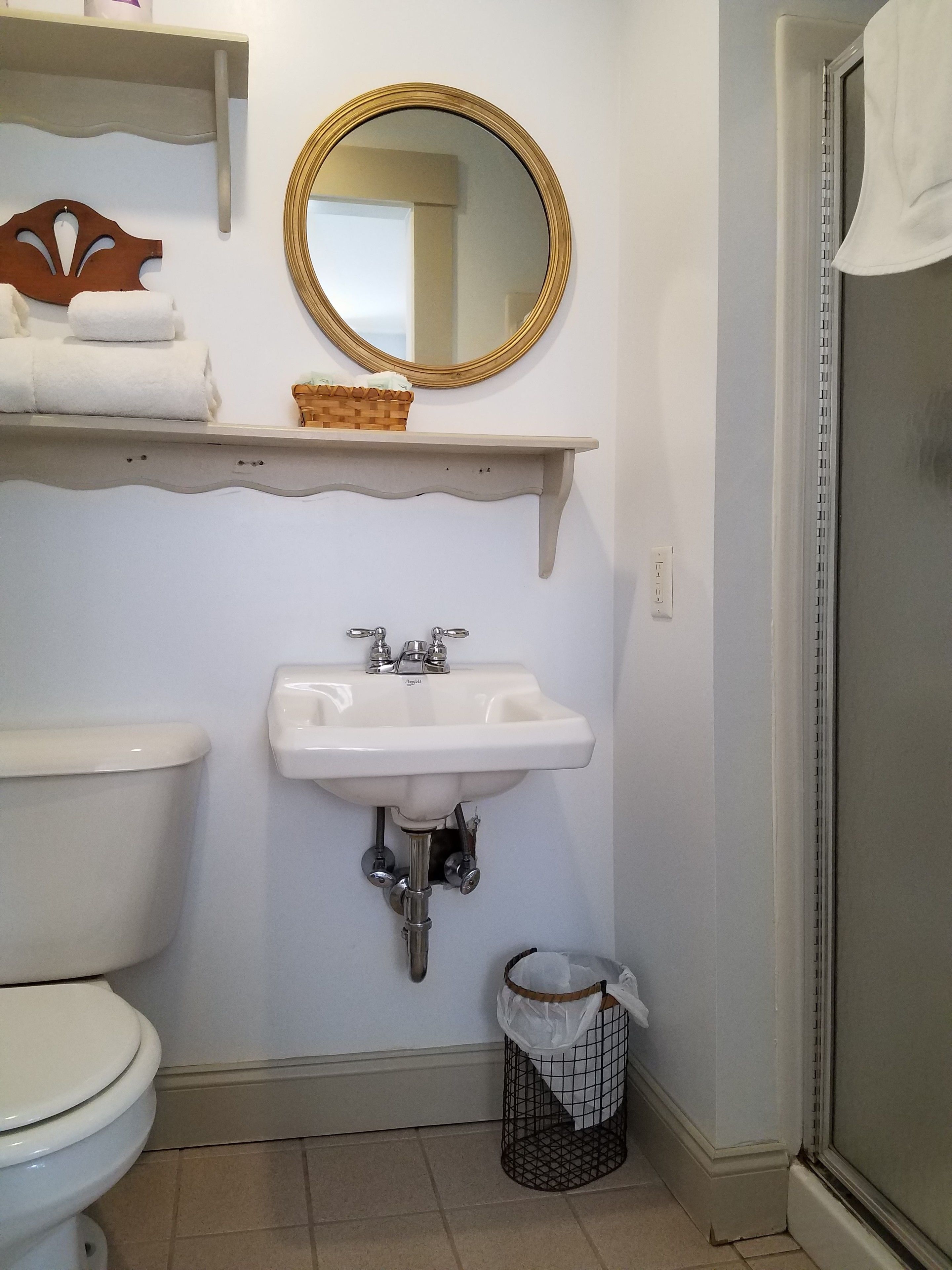 Une salle de bain avec des toilettes, un lavabo et un miroir. On y trouve des serviettes et un panier sur une étagère au-dessus du lavabo.