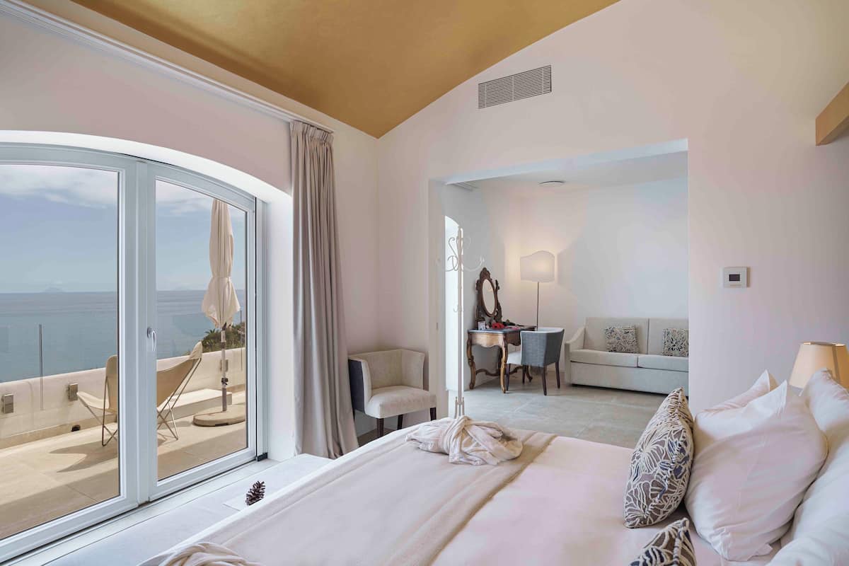 A hotel room with a large sliding glass door leading to a balcony, a bed with pillows, a desk with a chair, and a sofa in the background.