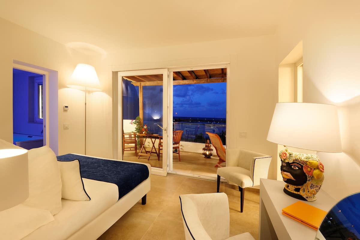 A hotel room with a bed, a sofa, a table with a lamp, and a balcony with a table and chairs.