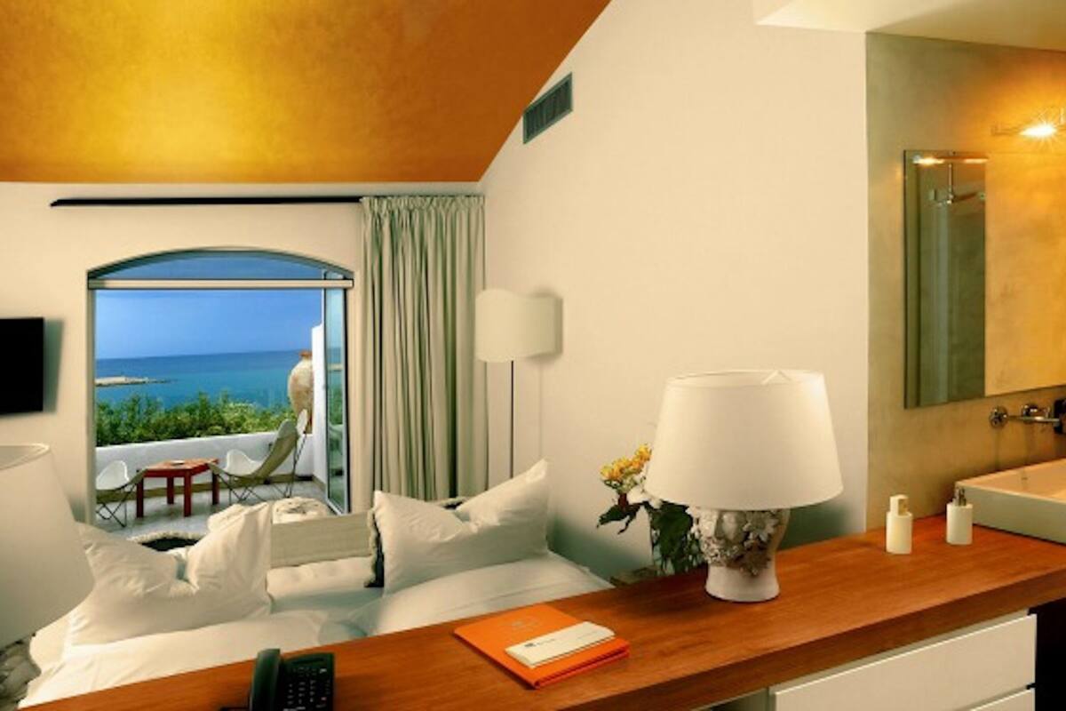 A hotel room with a bed, a desk, a lamp, a bathroom with a sink, and a view of the sea.