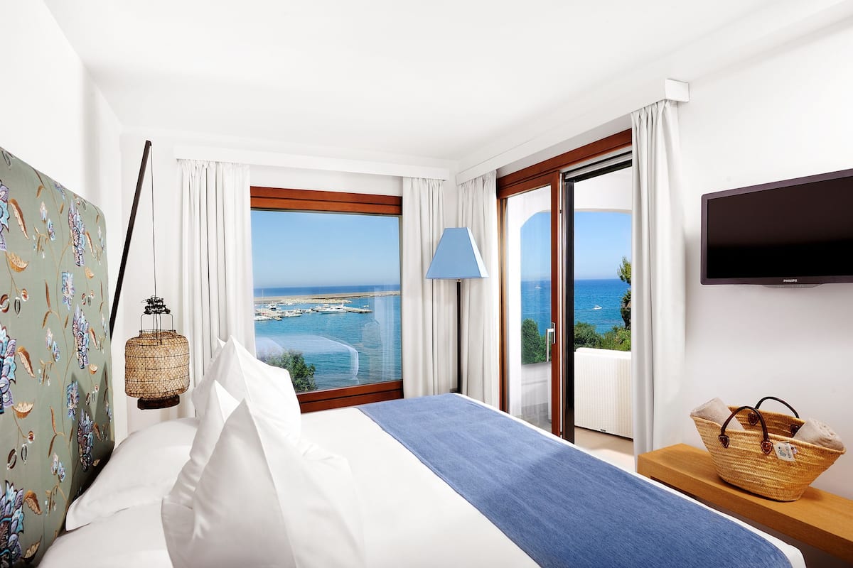 A hotel room with a bed, a television, a view of the sea, and a wicker basket on a nightstand.