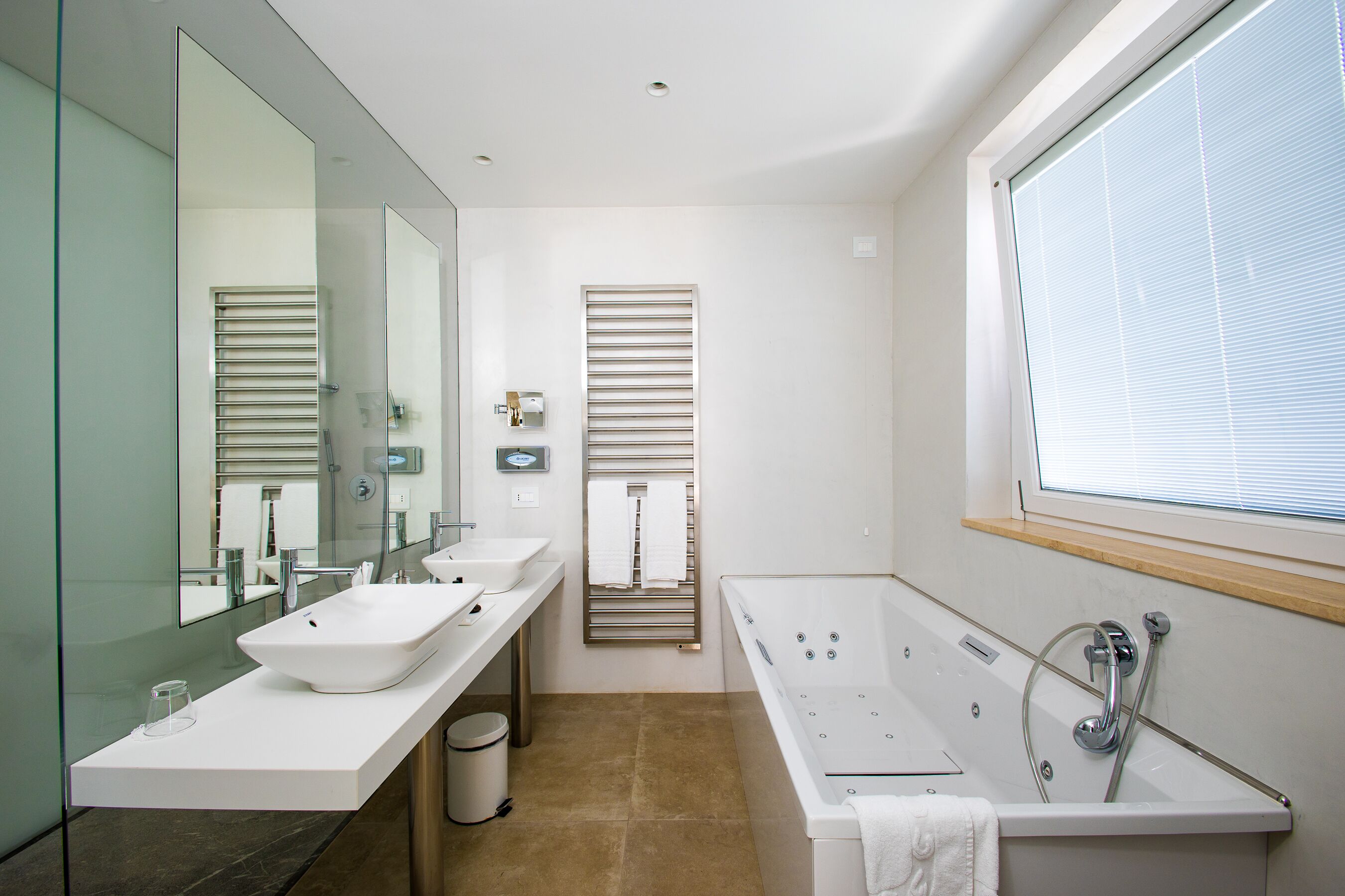 A modern bathroom with a large mirror, two sinks, a bathtub, and a window.