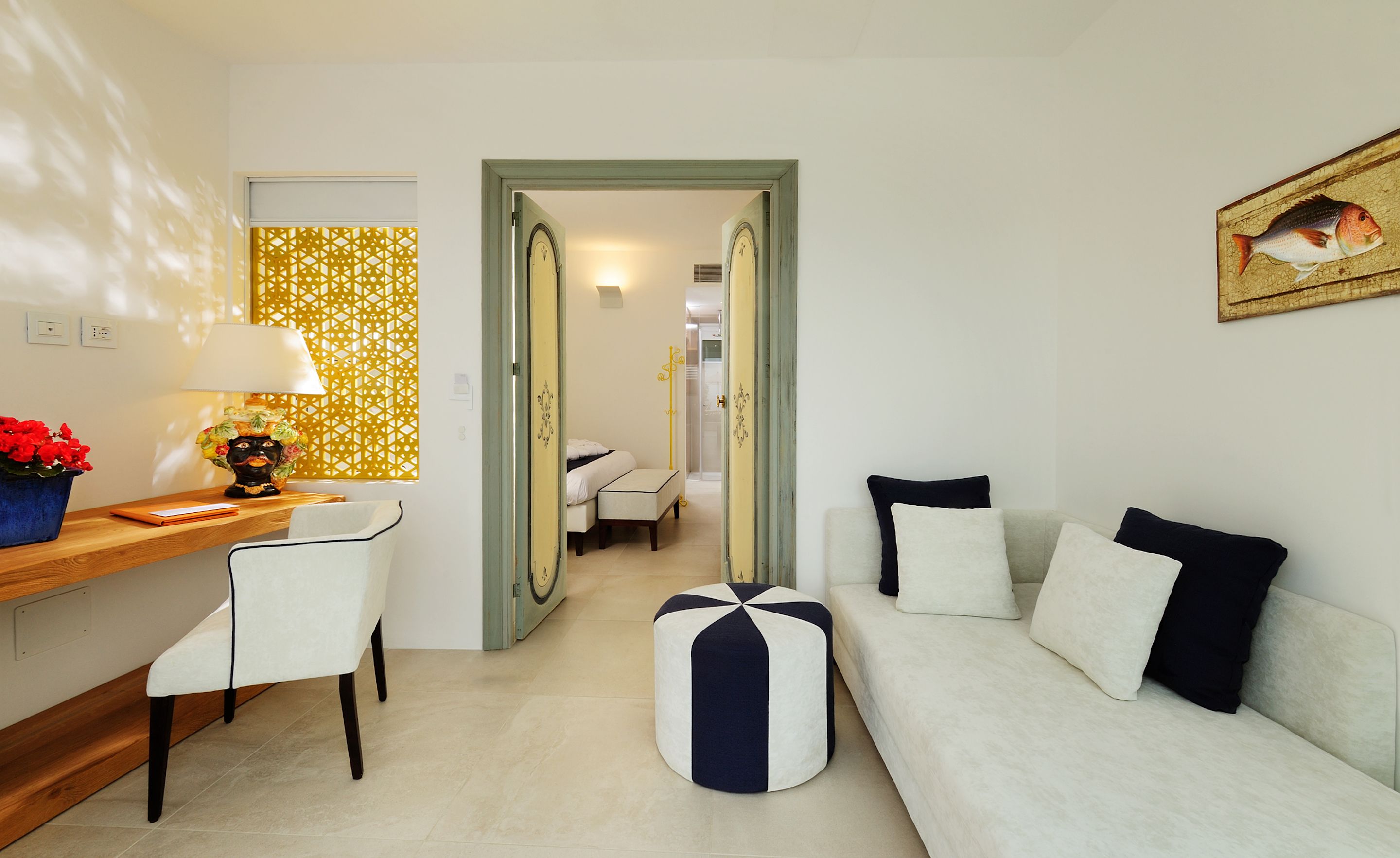 A modern hotel room with a wooden desk, a white sofa, a round ottoman, and a mirror on the wall.