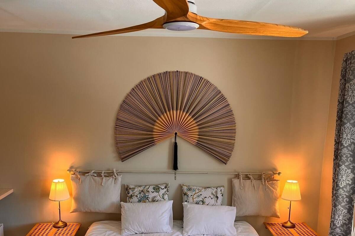 A hotel room with a bed, two bedside tables with lamps, a ceiling fan, and a wall-mounted decorative piece.
