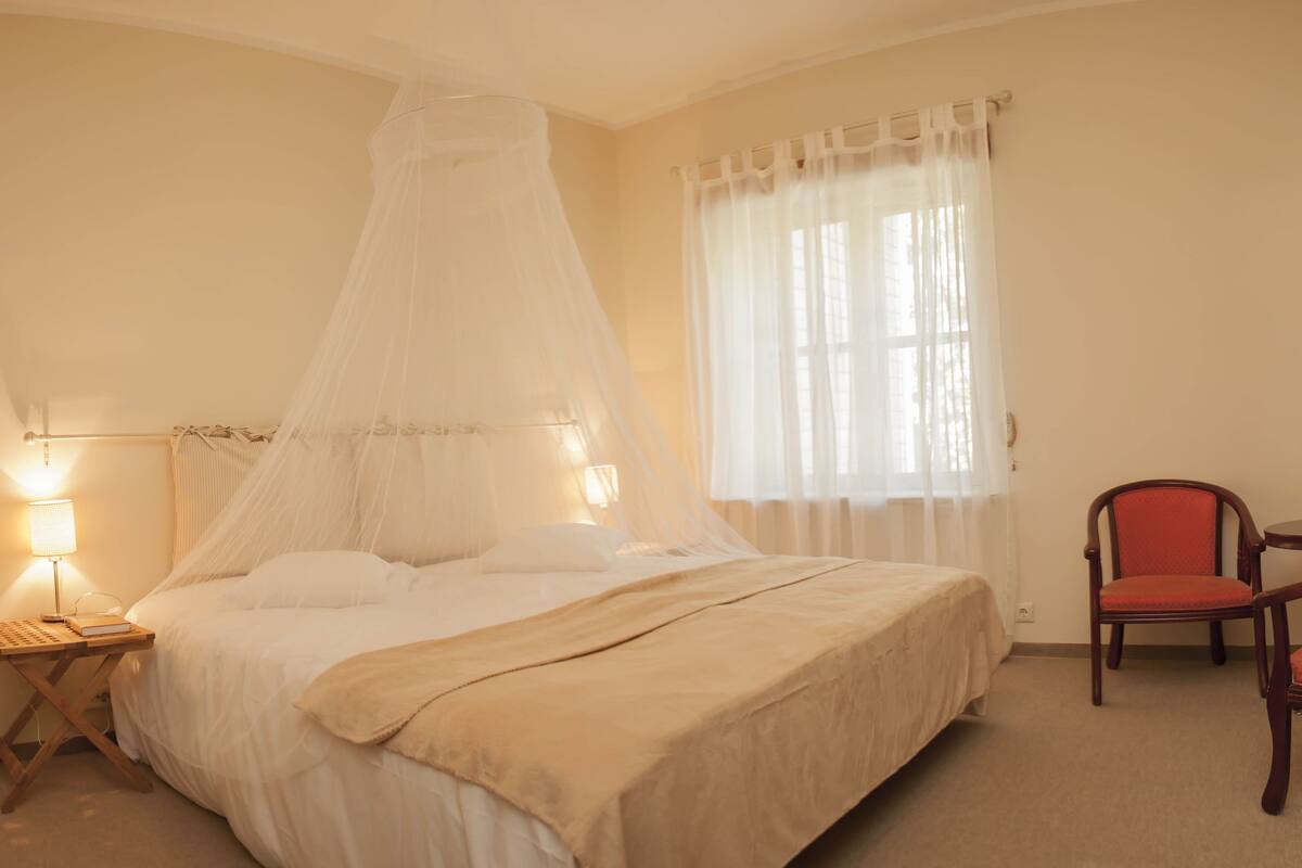 A hotel room with a bed, bedside table with a lamp, a chair, a small table, and a window with curtains.