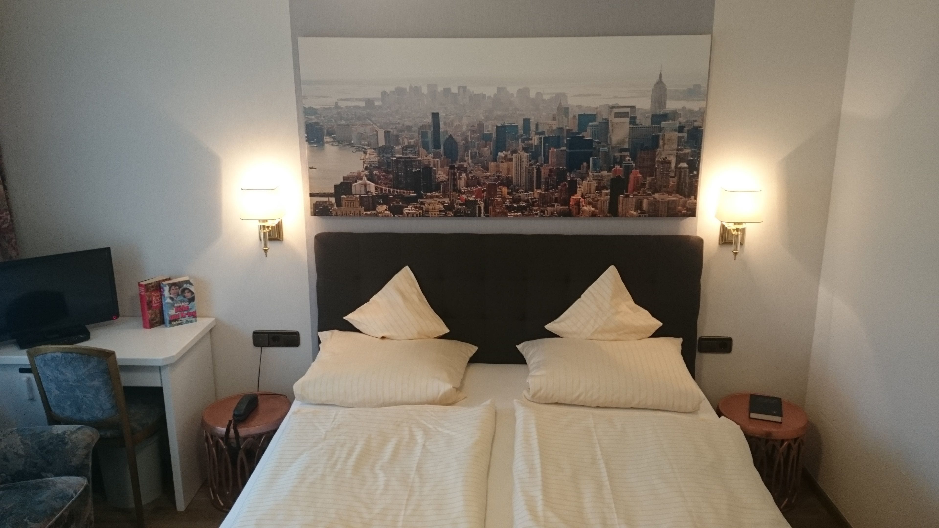A hotel room with a bed, two bedside tables, a desk with a computer, a chair, and a cityscape view on the wall.