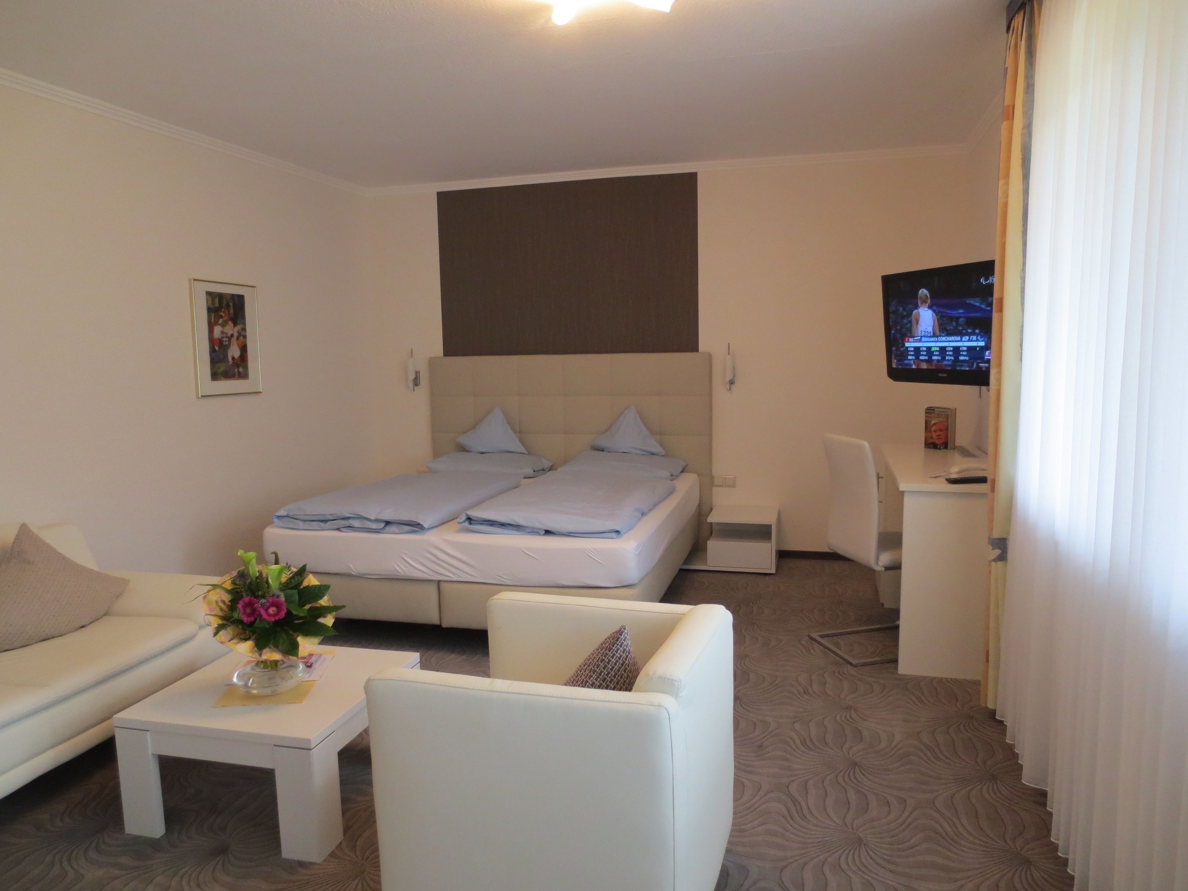 Una habitación de hotel con una cama, un sofá, una mesita con un florero y un escritorio con una computadora, además de un televisor en la pared.
