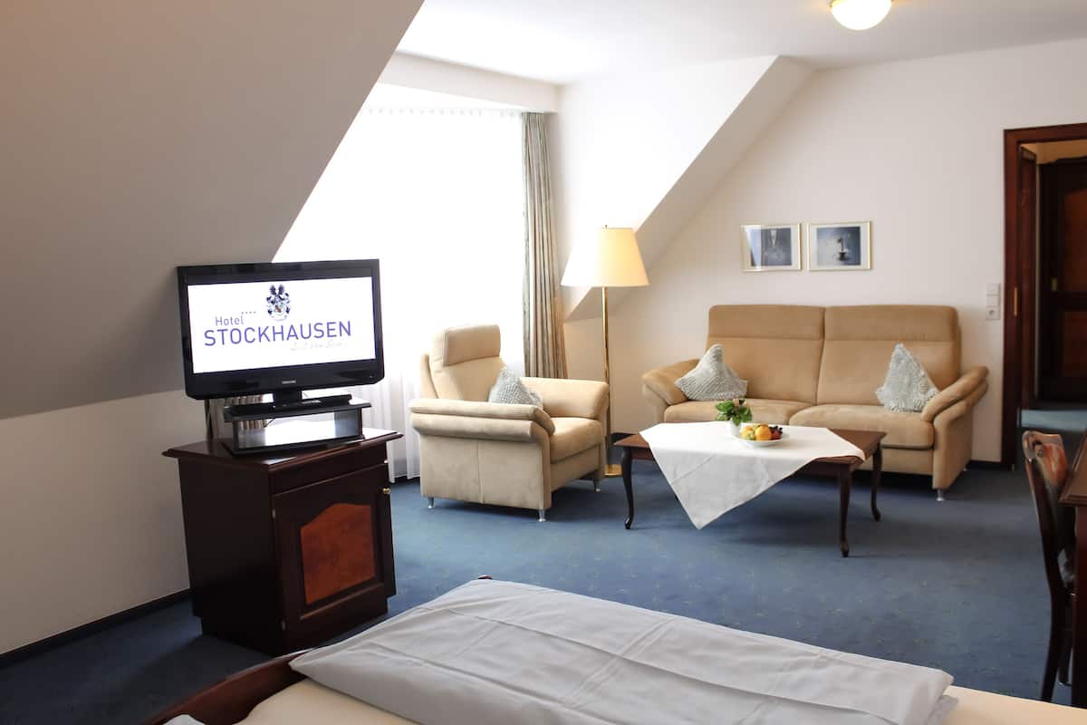 A hotel room with a TV displaying 'Hotel Stockhausen', a sofa, armchairs, a small table with a fruit bowl, and a bed.