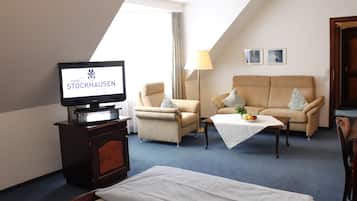 Una habitación de hotel con una televisión mostrando 'Hotel Stockhausen', un sofá, sillones, una mesita con un frutero y una cama.