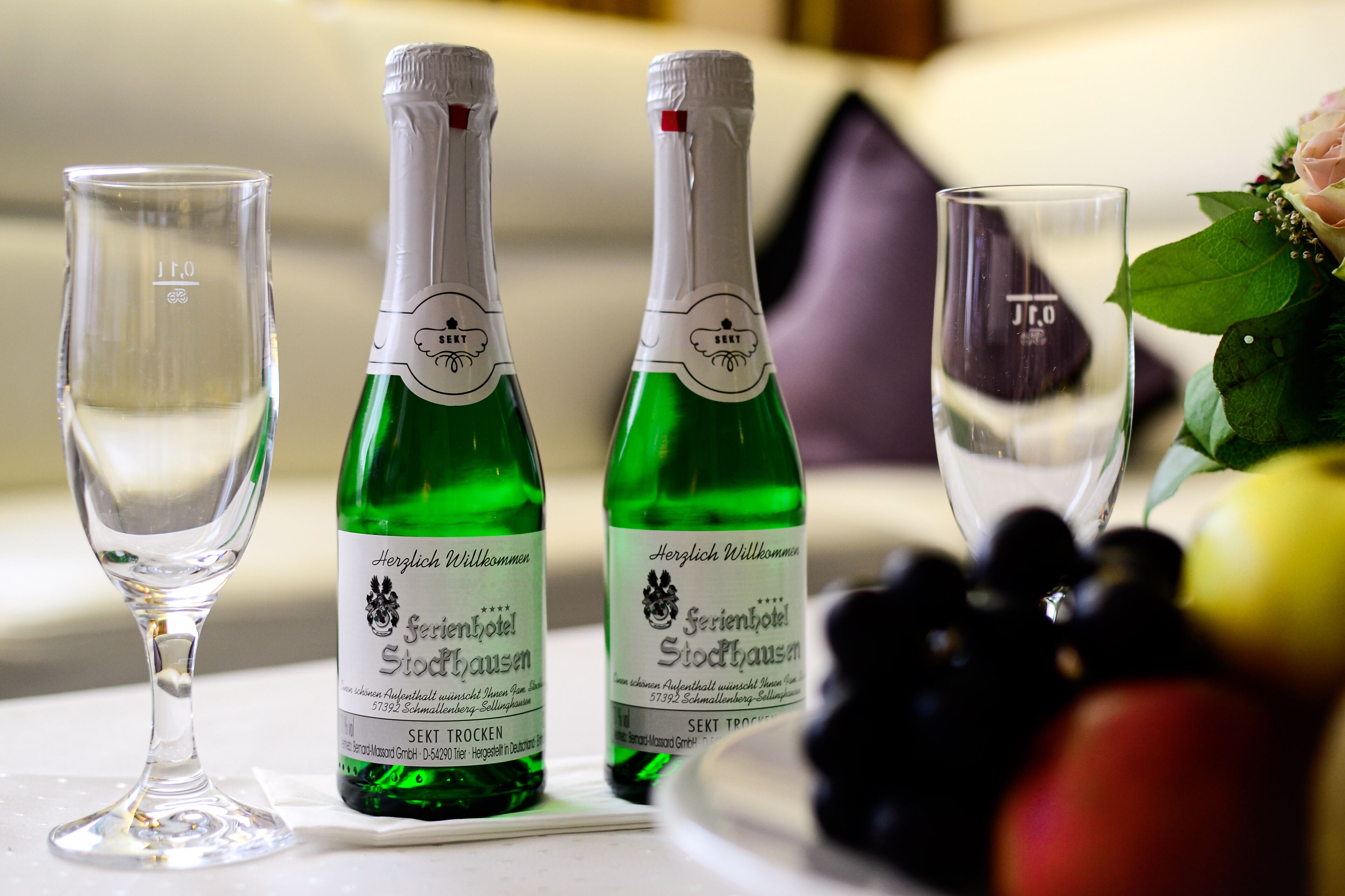 Two green bottles of Sekt Trocken next to three clear glasses on a table, with a bowl of grapes and a bouquet of flowers in the background.