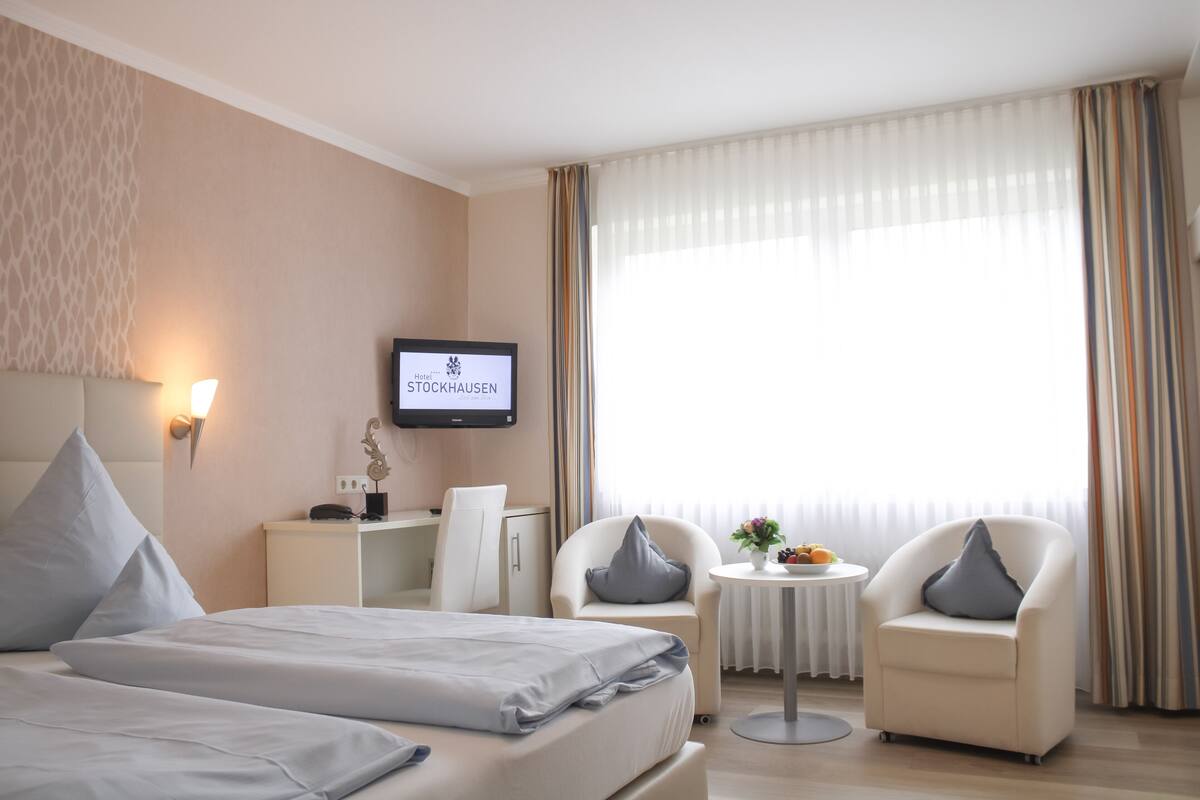 A hotel room with a bed, two armchairs, a small table with a vase and fruit, a TV on the wall, and a window with curtains.