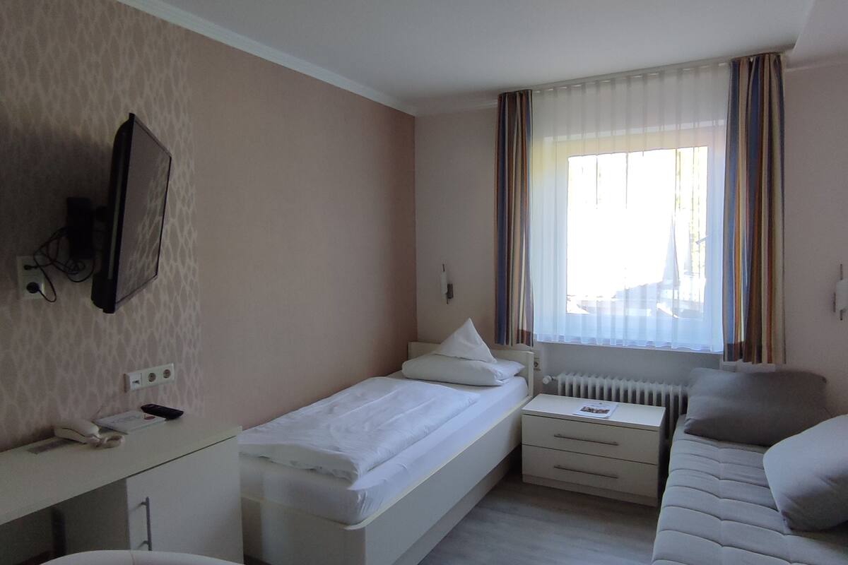 A bedroom with a single bed, a desk, a chair, a sofa, a TV mounted on the wall, and a window with curtains.
