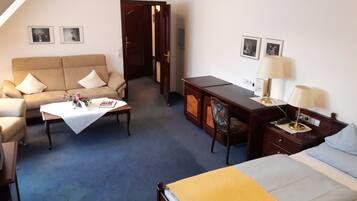 Quarto de hotel com cama, sofá, escrivaninha e cadeira. Há duas luminárias na escrivaninha e uma porta que dá para outro cômodo.