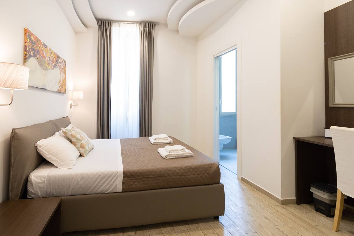 A modern hotel room with a large bed, a desk, and a bathroom visible through an open door.