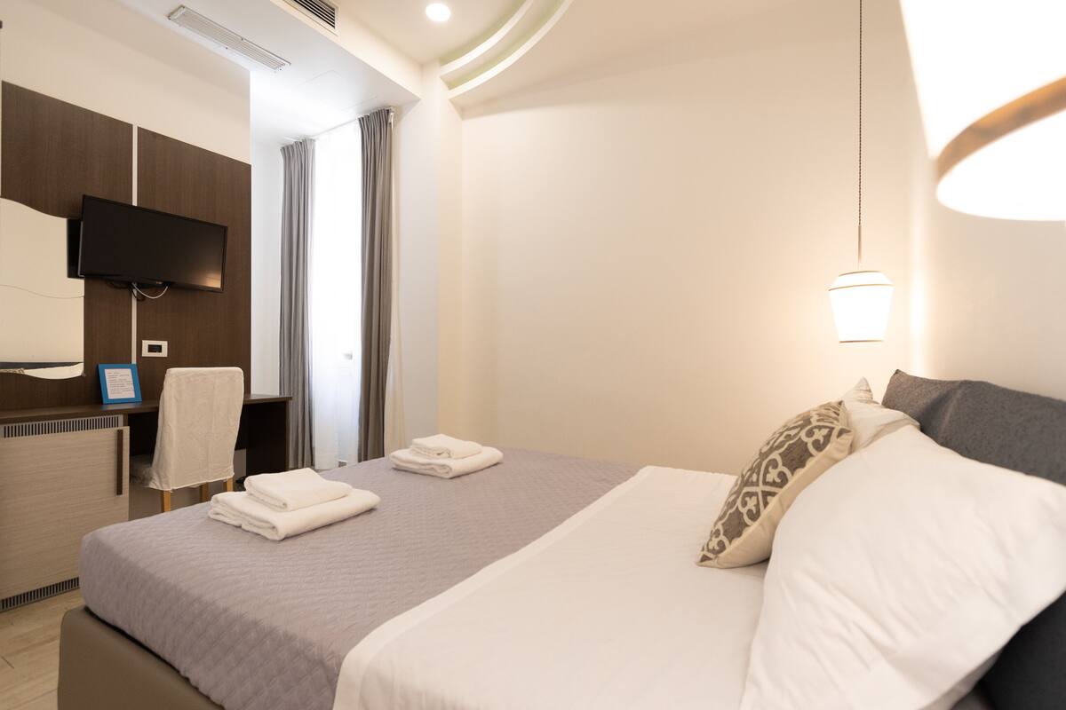 A hotel room with a bed, a desk, a flat-screen TV, and a wall-mounted light fixture.