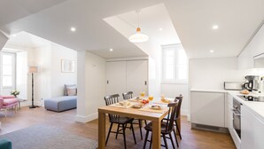 A modern kitchen with a dining table, chairs, and a breakfast setup. There is a sofa and a lamp in the adjacent living area. - LSA Bairro Alto by Numa (Lisbon)