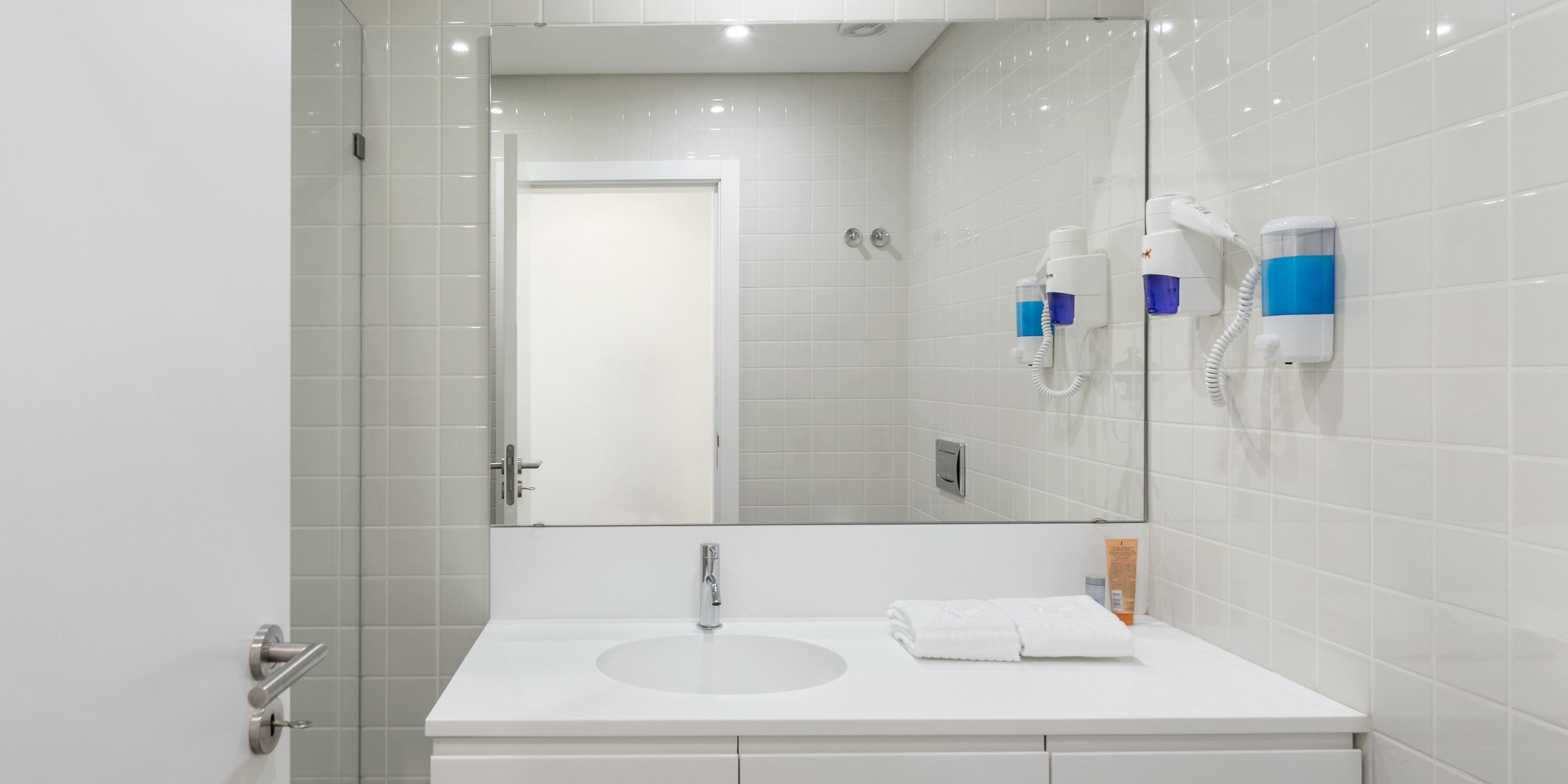 Une salle de bain moderne avec un lavabo blanc, un miroir et deux sèche-cheveux fixés au mur.